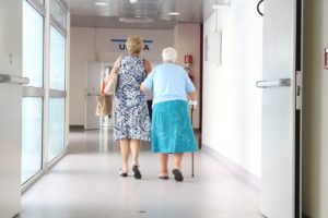 an elderly woman walks down a hospital hall supported by another woman